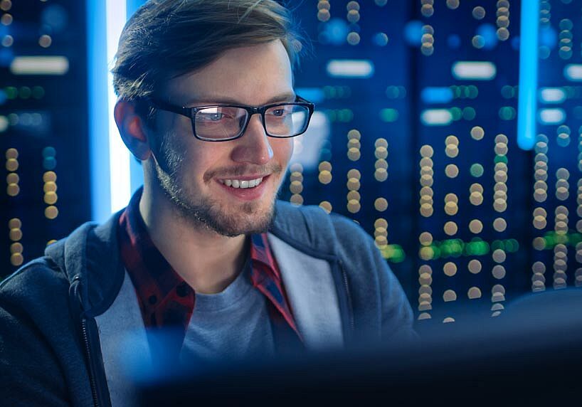 Experte für Systemhärtung | Mann mit Brille sitzt vor einem Computer, lacht und im Hintergrund sind Lichter von Servern zu sehen
